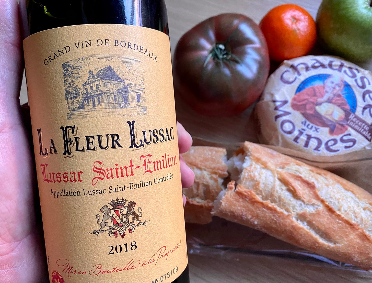 A bottle of La Fleur Lussac Saint emission 2018 red wine in the foreground and a baguette, Chaussee aux Moines cheese and a large beef tomato.