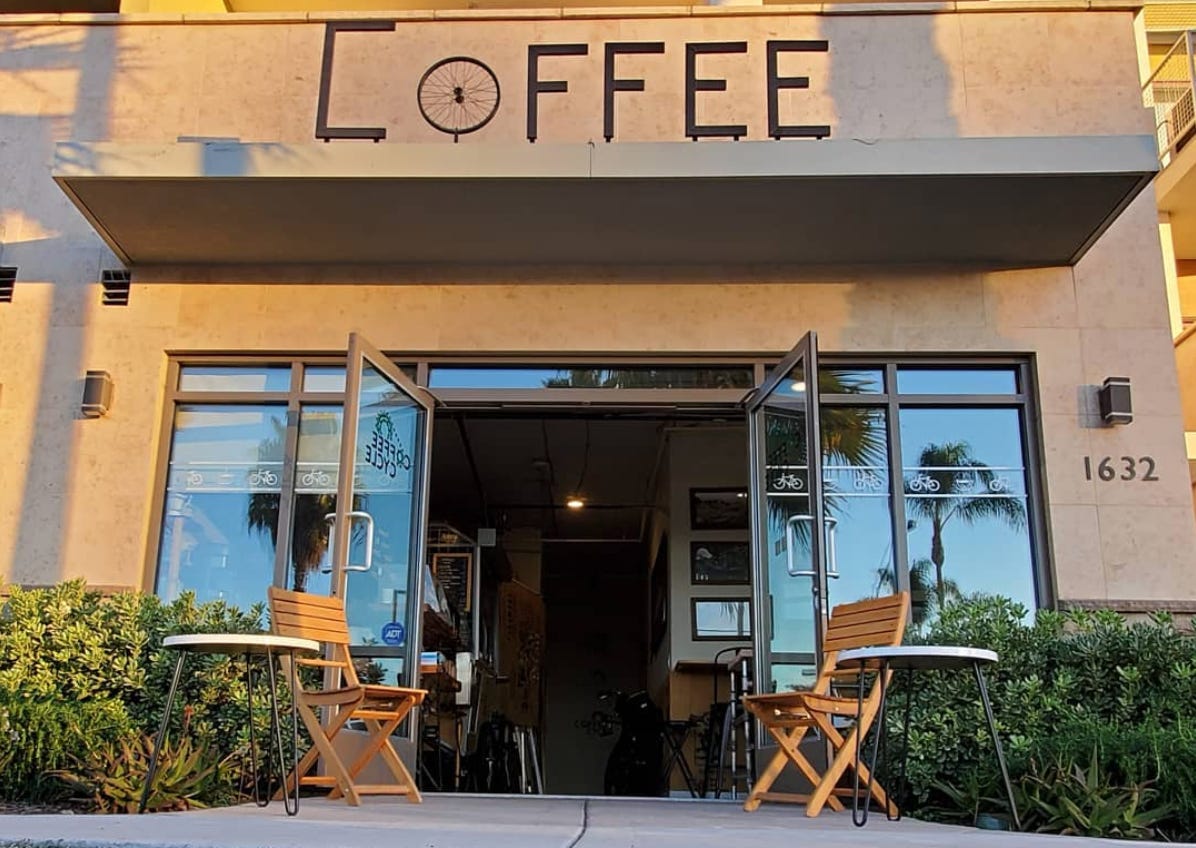 A photo from a low angle of the entry way to Coffee Cycle cafe in Pacific Beach, San Diego, California. A chair and side table frame the open doorways and the word coffee in black iron wrought letters is mounted above the door.