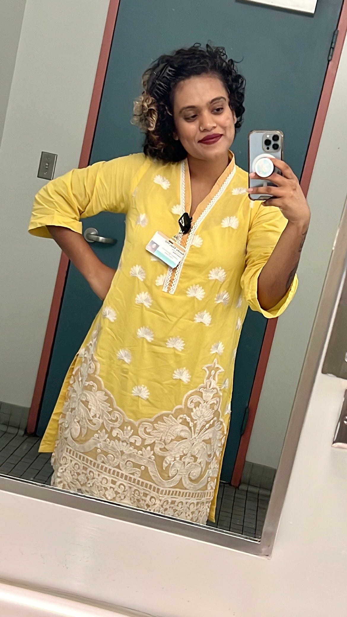 Ayesha taking a selfie in a bathroom mirror at work wearing a handmade, vibrant, yellow, traditional South Asian Shalwar Kameez outfit