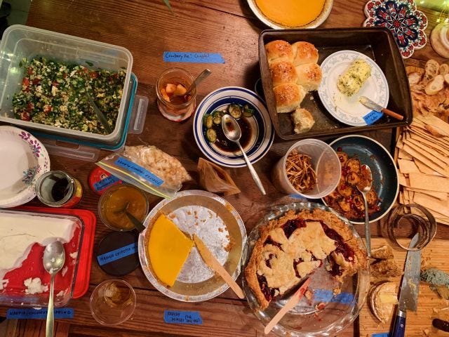 A table crowded with many dishes labeled with blue tape, from rolls to pies to salads