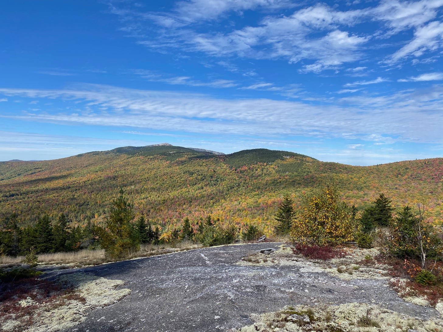 Vista of a mountain ridge under a bright blue sky dotted with clouds. The forested mountain slopes are a mixture of green, red, orange, and gold.