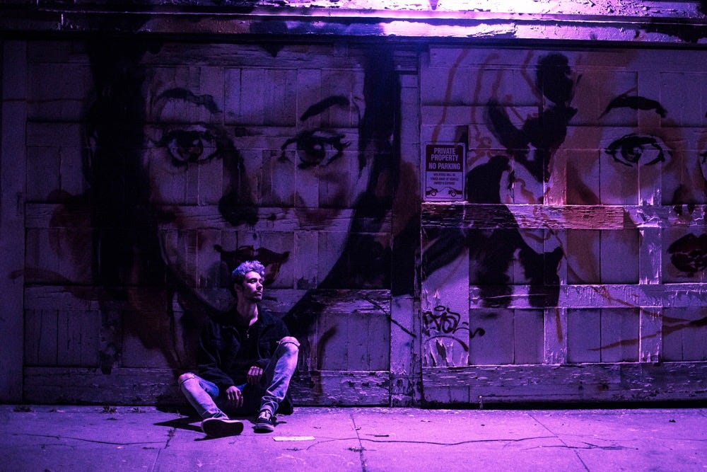  cøzybøy sitting on the ground outside in front of a mural of a portrait of a woman with purple and pink lighting.
