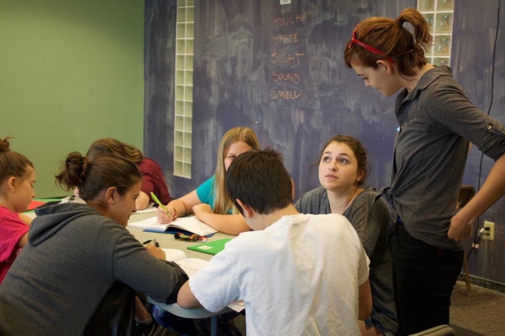 Austin Bat Cave students sitting at a white rectangle table, writing and discussing their work with an ABC instructor who is standing by the table near a blackboard in the background. Austin Bat Cave students sitting at a white rectangle table, writing and discussing their work with an ABC instructor who is standing by the table near a blackboard in the background.