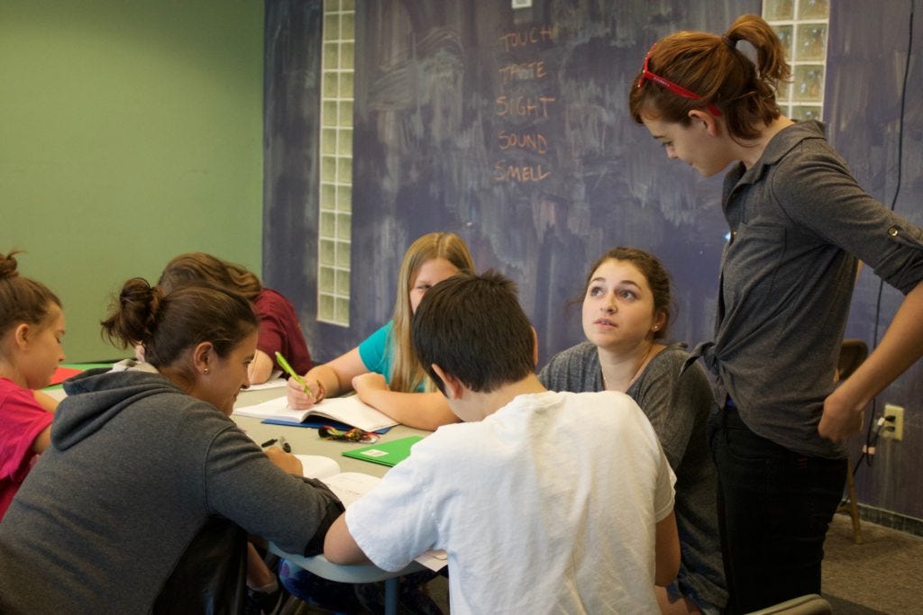 Austin Bat Cave students sitting at a white rectangle table, writing and discussing their work with an ABC instructor who is standing by the table near a blackboard in the background. 