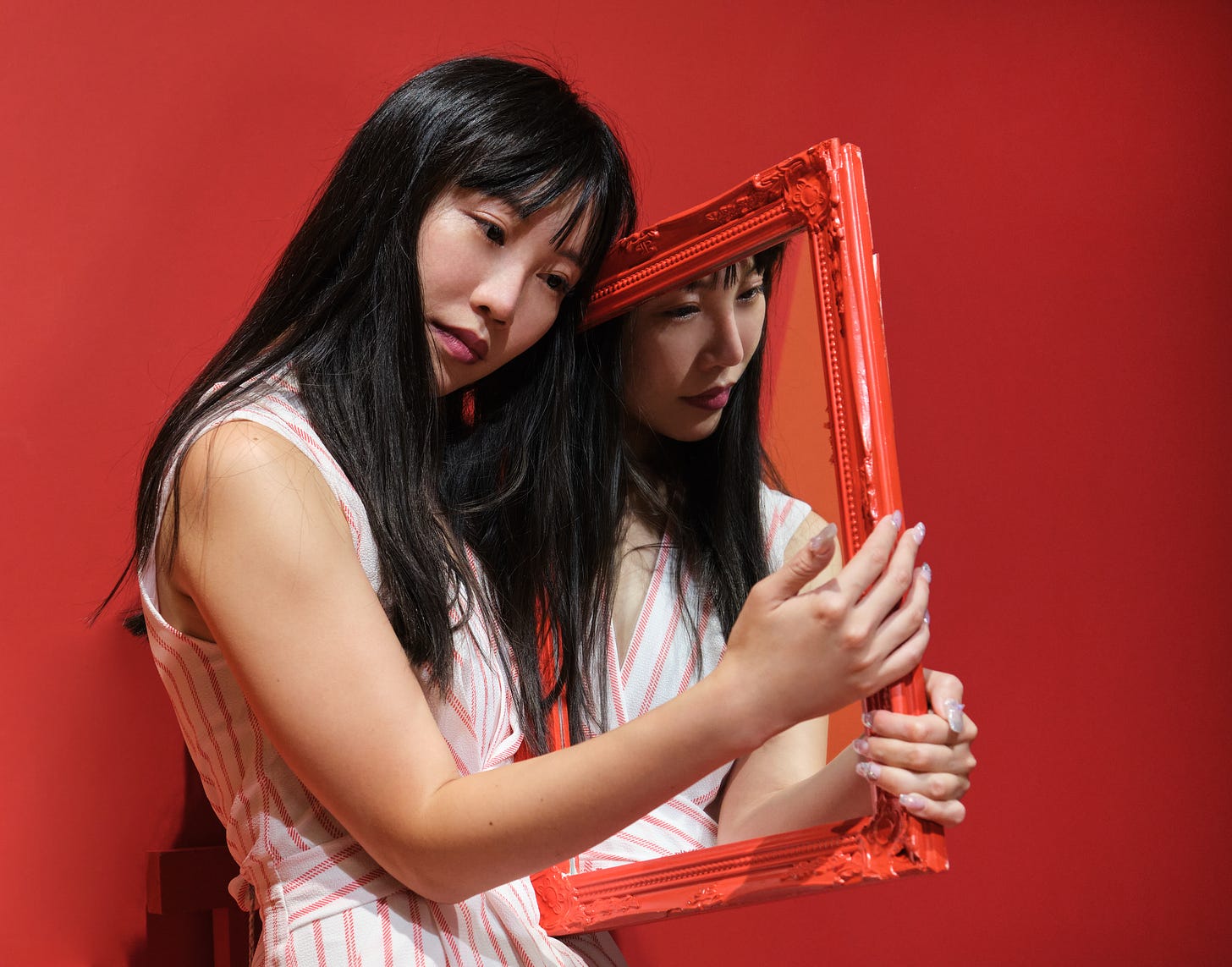 Young asian woman pose with a red framed mirror over a red background. Her reflection emerges from one side to help hold the mirror.