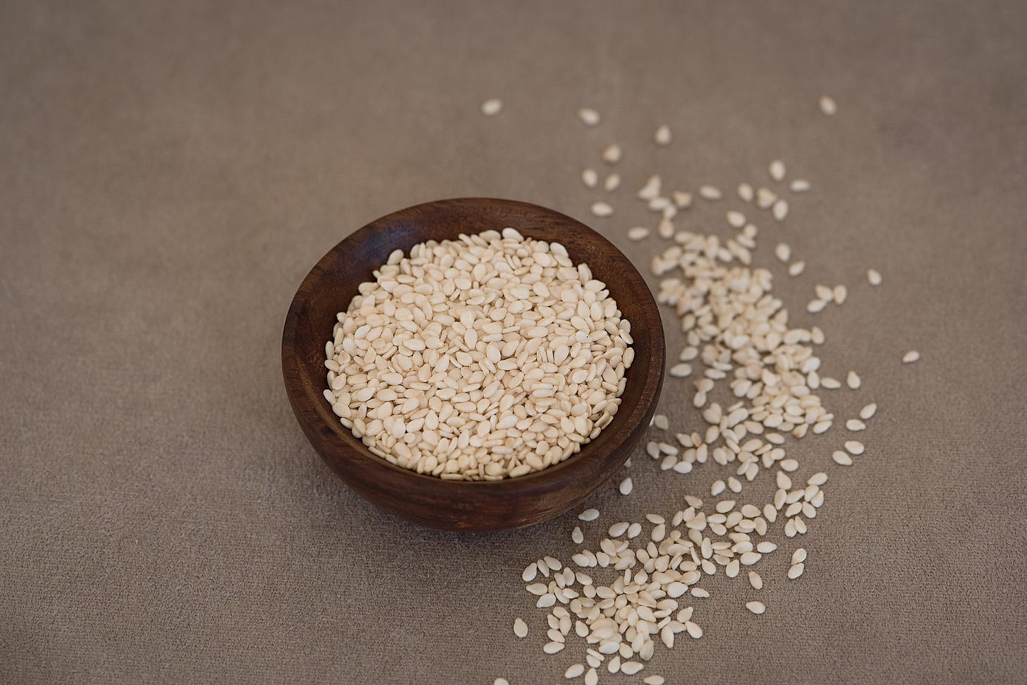 A bowl containing sesame seeds, there are also seeds spread in an arch on the right side.