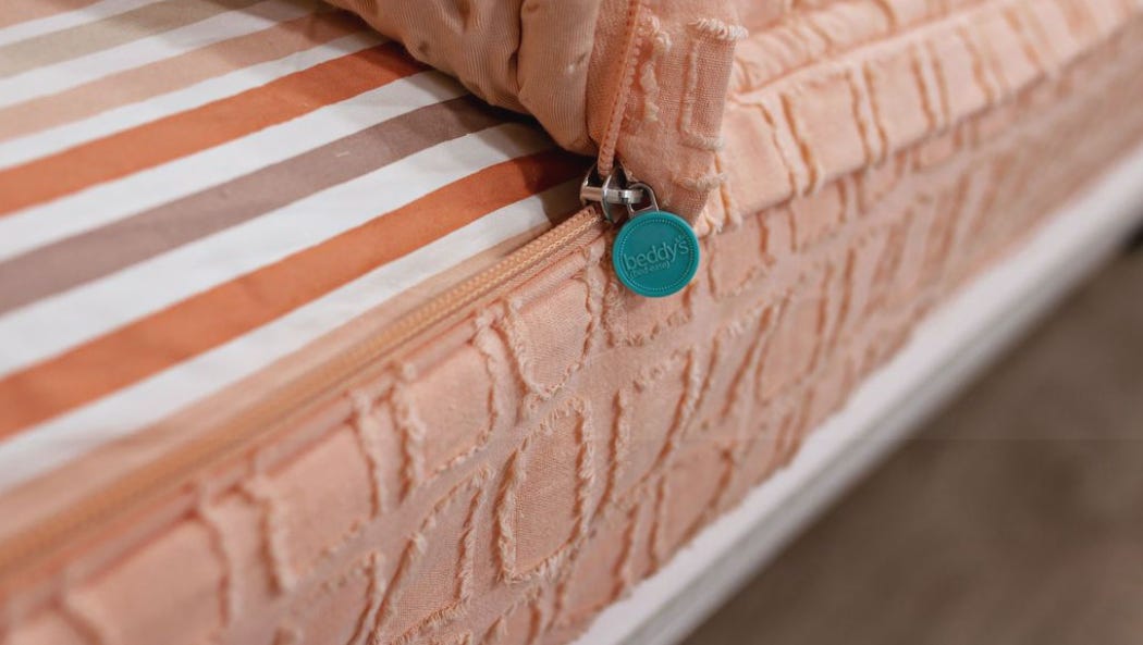 An orangish-pind comforter tucked into a bed. It has a zipper cutting through the edge of it so someone can slip into the bed under it without removing it. There is a fitted sheet as well as a minky textured blanket within the zipped comforter.