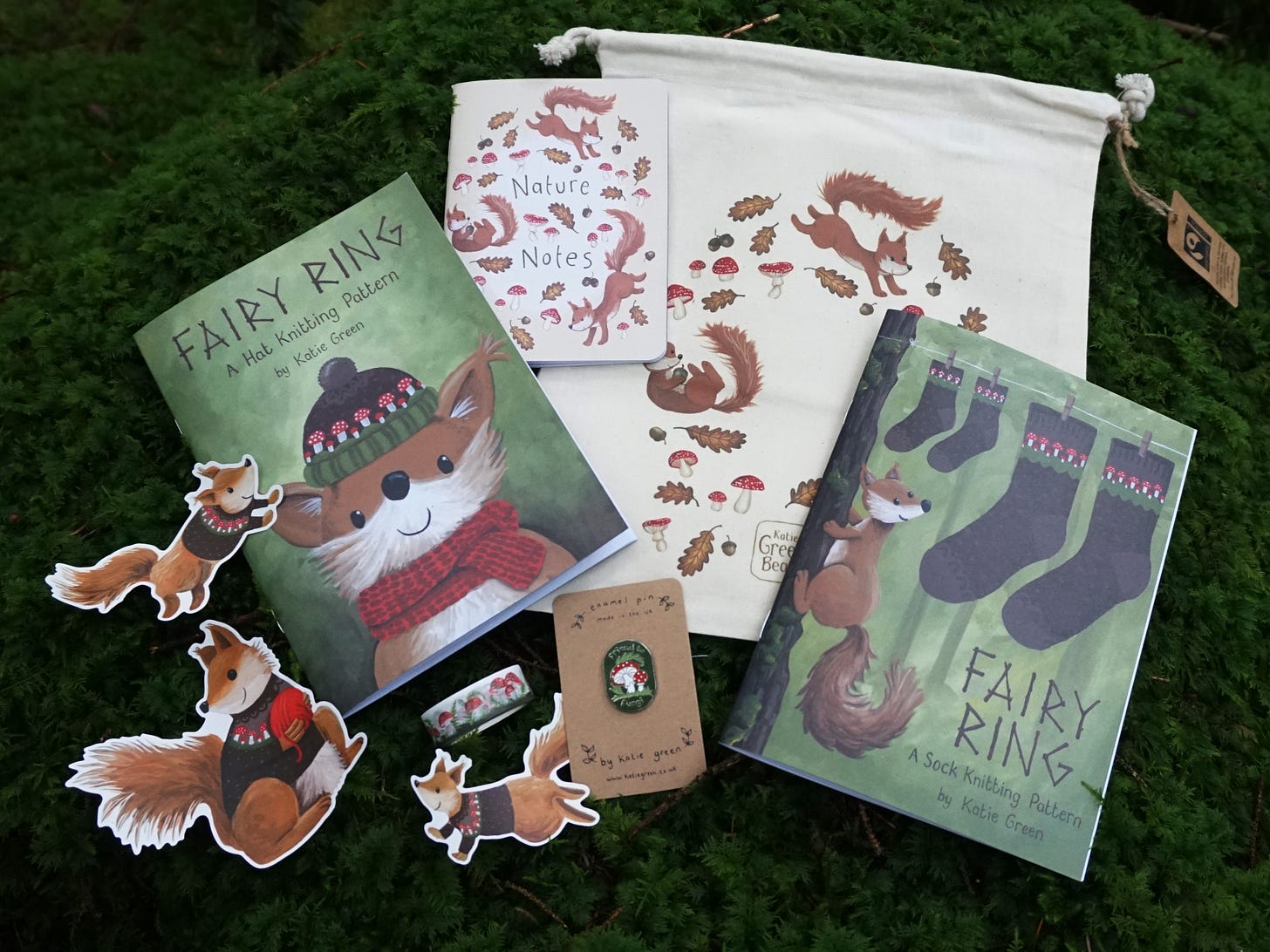 Image description: A pile of cute goods laid out on a bed of squishy green moss. There is an unbleached cotton drawstring project bag printed with tumbling squirrels, leaves and mushrooms. There's a little notebook entitled Nature Notes with the same squirrl illustration on the front. There are two illustrated pattern booklets, three cute illustrated stickers of squirrels wearing cardigans, a roll of mushroom washi tape and an enamel pin.