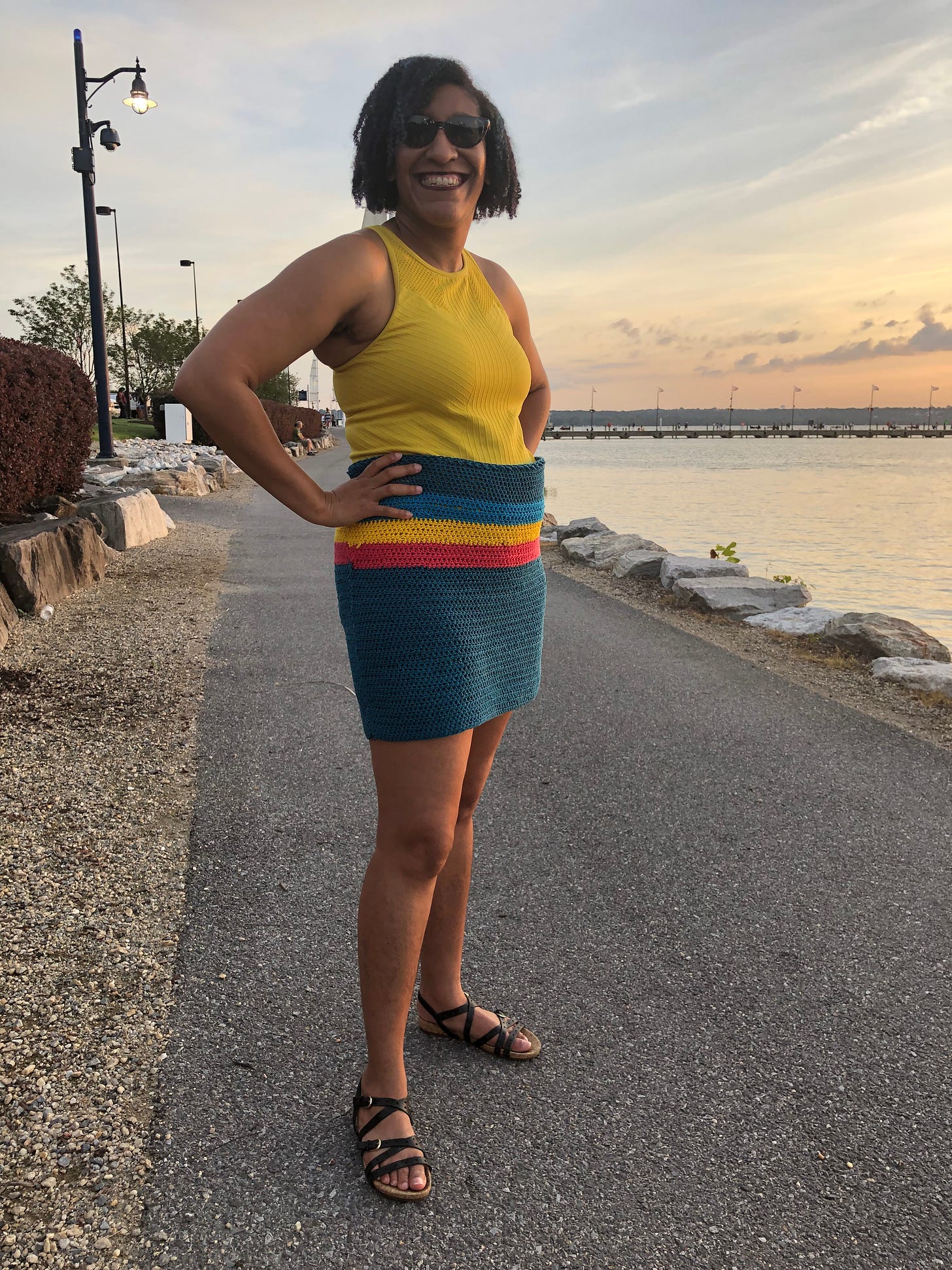 Kristen is standing with her hands-on her hips on her self crocheted rainbow skirt and with a rainbow tank top on the Potomac River trail portion of National Harbor