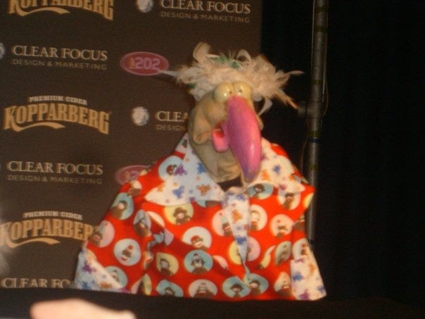 A photograph of a cartoonish turkey puppet standing behind a podium. He is wearing a red suit with pictures of sock monkeys on it and has absurd tousled hair. 