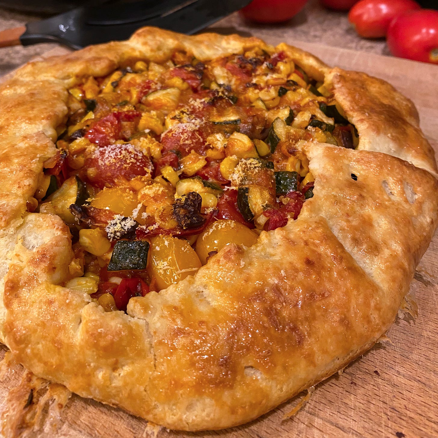A golden brown galette on a cutting board, filled with a mix of orange and red grape tomatoes, pieces of corn, zucchini, and olives. Grated parmesan lightly covers the top.