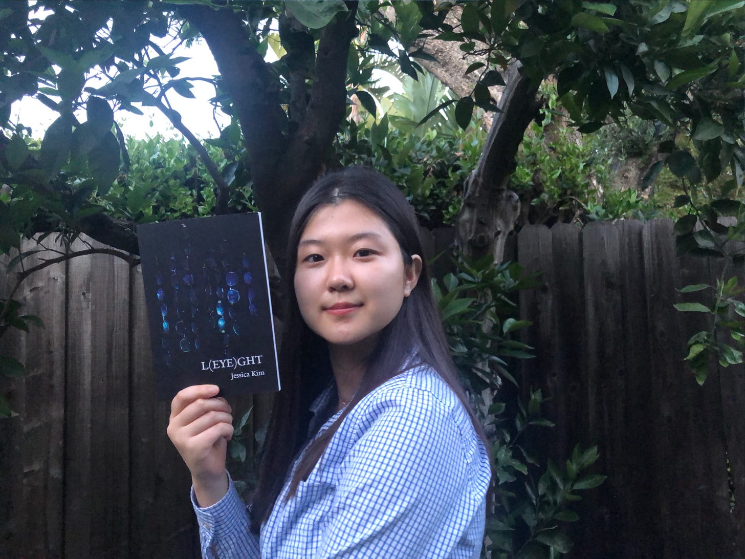 Jessica, a Korean-American, is wearing a blue blouse with tiny blue squares all over it. Not sure what you call that. She is holding her book up to the camera. She is standing outside in a fenced backyard, likely being bit by mosquitoes.