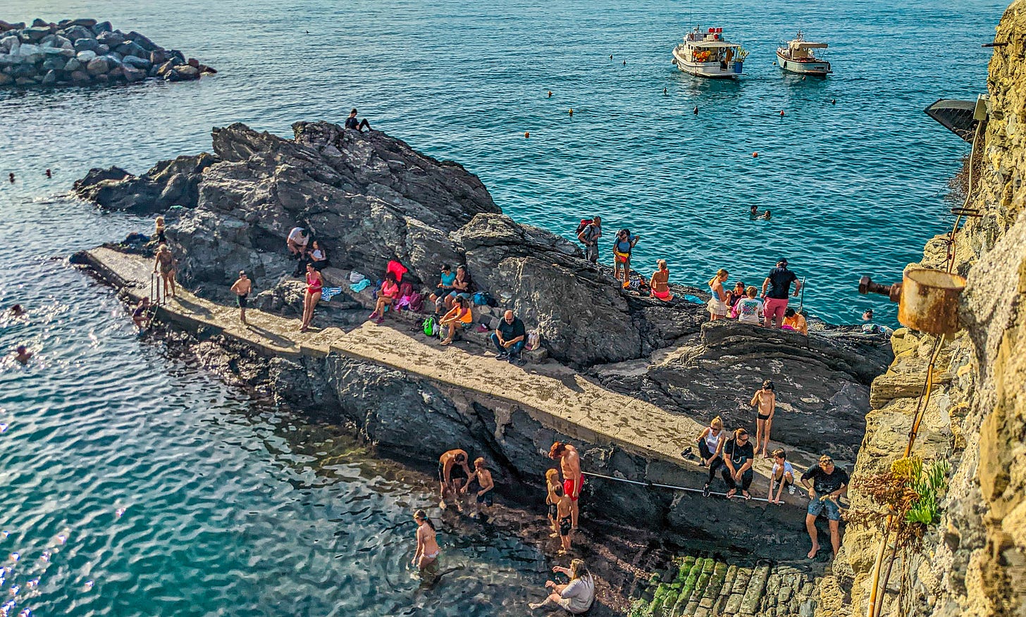 A rock juts out into the blue harbor, swimmers in the water while others sit on the rock. 