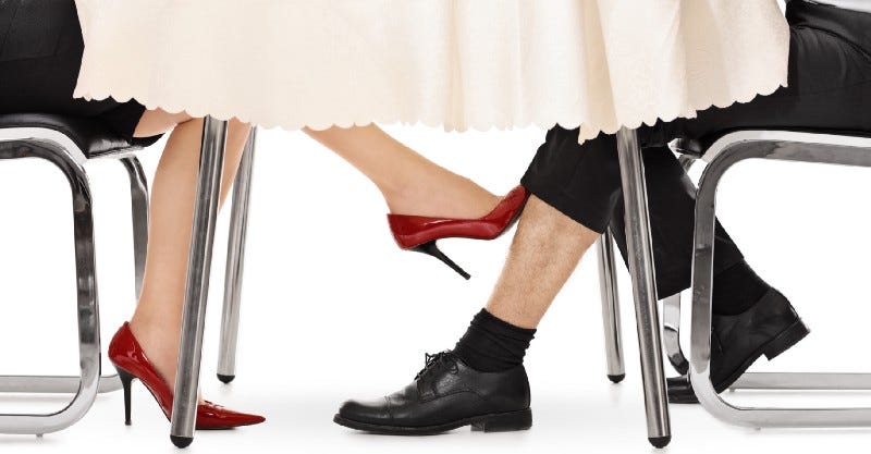 Under a table, a woman's foot in red high heels touches the leg of a man in dark slacks and dress shoes.