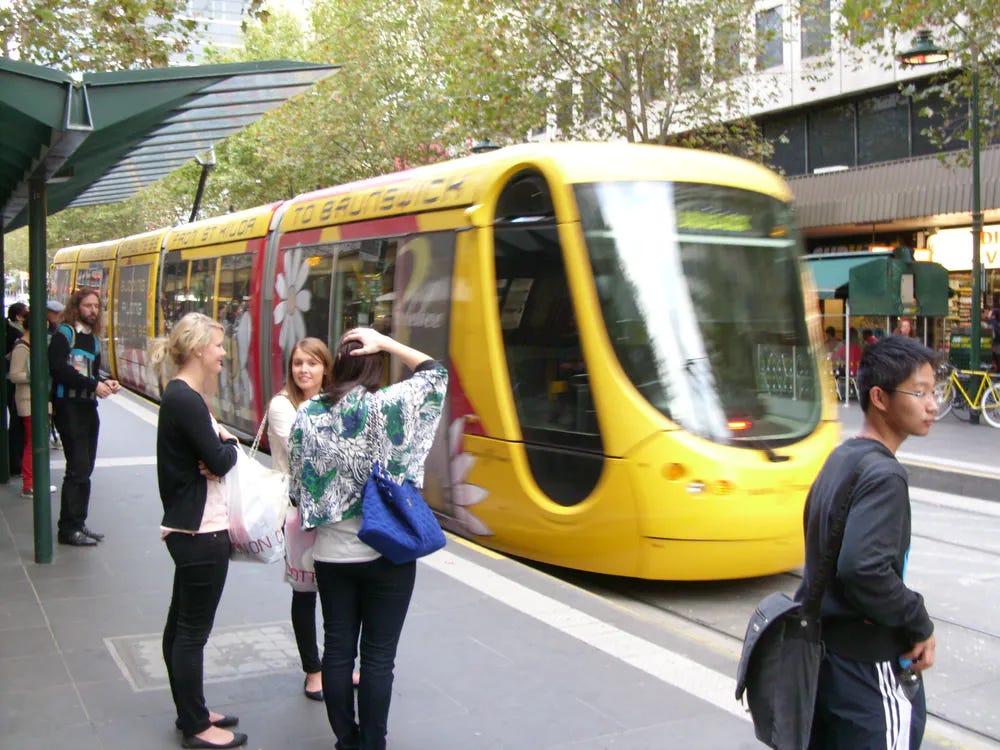 A yellow tram is leaving a stop in Melbourne, Australia. Three women are on the platform in conversation, while a young Asian man looks off to the right. There is a green canopy on the platform and trees in the background