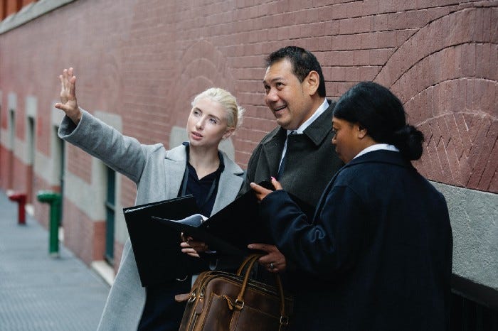 3 people standing on the sidewalk, two women, one male. Both women are talking to the guy in the center, who is holding a folder with some sort of plan. The woman on the left is gesturing towards something to explain something, and the woman on the right is pointing to something in the plan to give emphasis. The man in the center is smiling while looking at what the woman is pointing at.