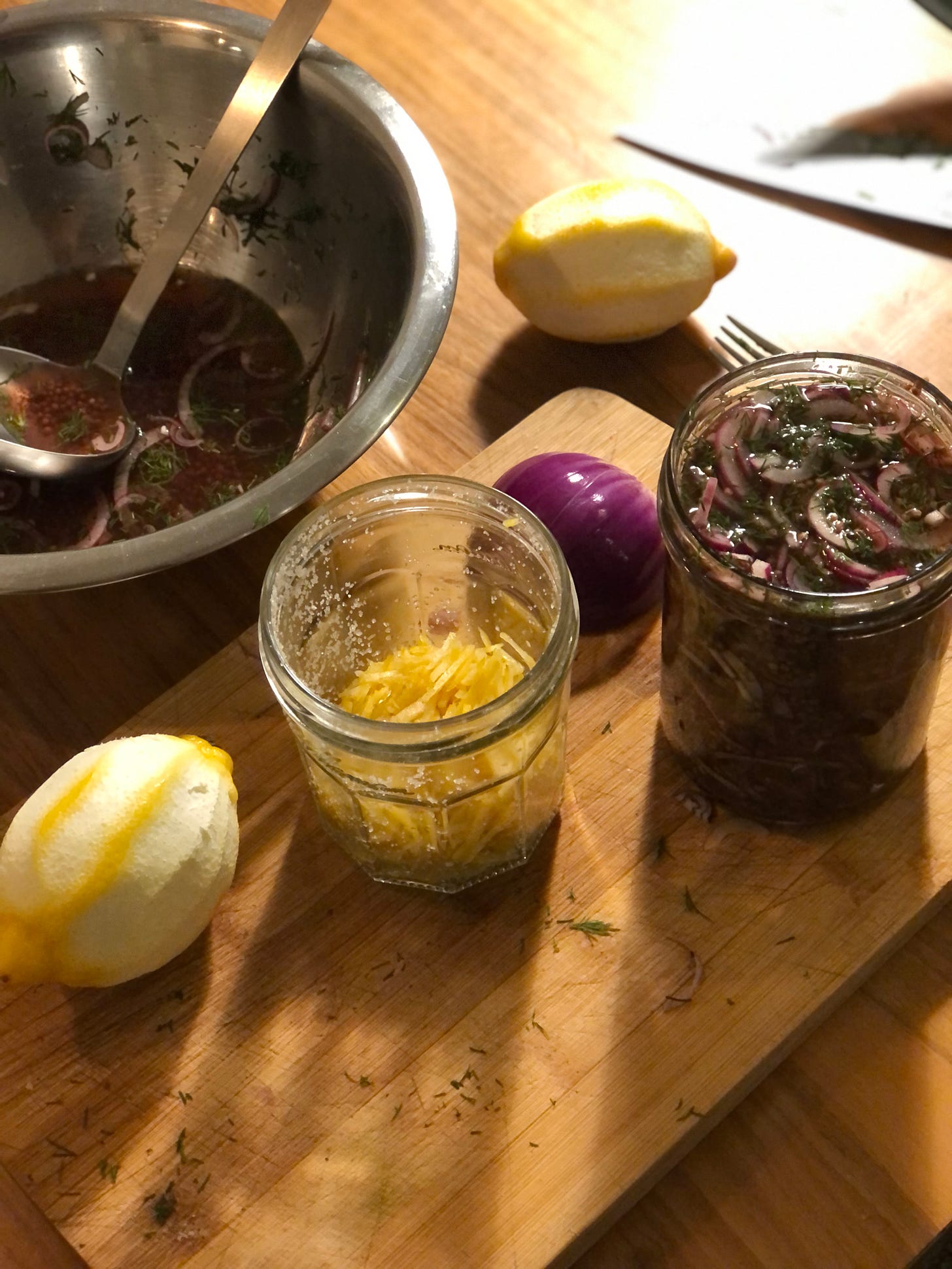 An angled overhead photo of a wooden table, clockwise from top left: a metal bowl with ladle and a pinky liquid with dill and onion slices; a lemon on its side with rind peeled off; on a wooden chopping board with a full jar of red onion slices in liquid, half a red onion, another glass jar half full with pieces of rind, another lemon.