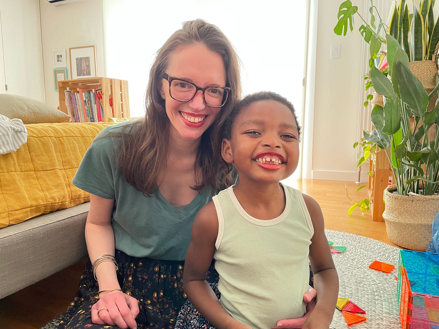 A white woman with long brown hair (Jessica) sits with a Black kid (Khalil) both are grinning and wearing bright red lipstick. 