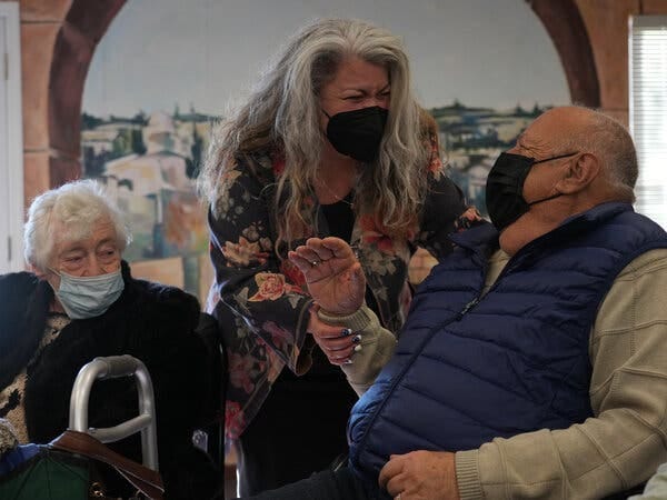 Debbie Toth wears a black face mask and shares a warm moment with Alexandr Makedonsky, who also wears a mask and sits in a chair wearing a blue vest and a tan-green sweater. Ms. Toth holds Mr. Makedonsky’s wrist, and to their left, Tsilya Tankover wears a face mask and a large dark coat draped over her sweater.