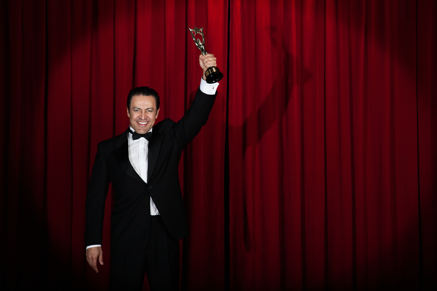 Man in tux on stage in front of red curtain holding Emmy statuette aloft with his left hand