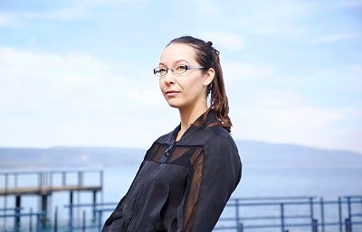 Picture of Essa Hansen. She has brown hair tied back and glasses and is looking diagonally past the camera. She is wearing a blue and brown jacket and standing outdoors, with a body of water and a blue sky visible in the background.