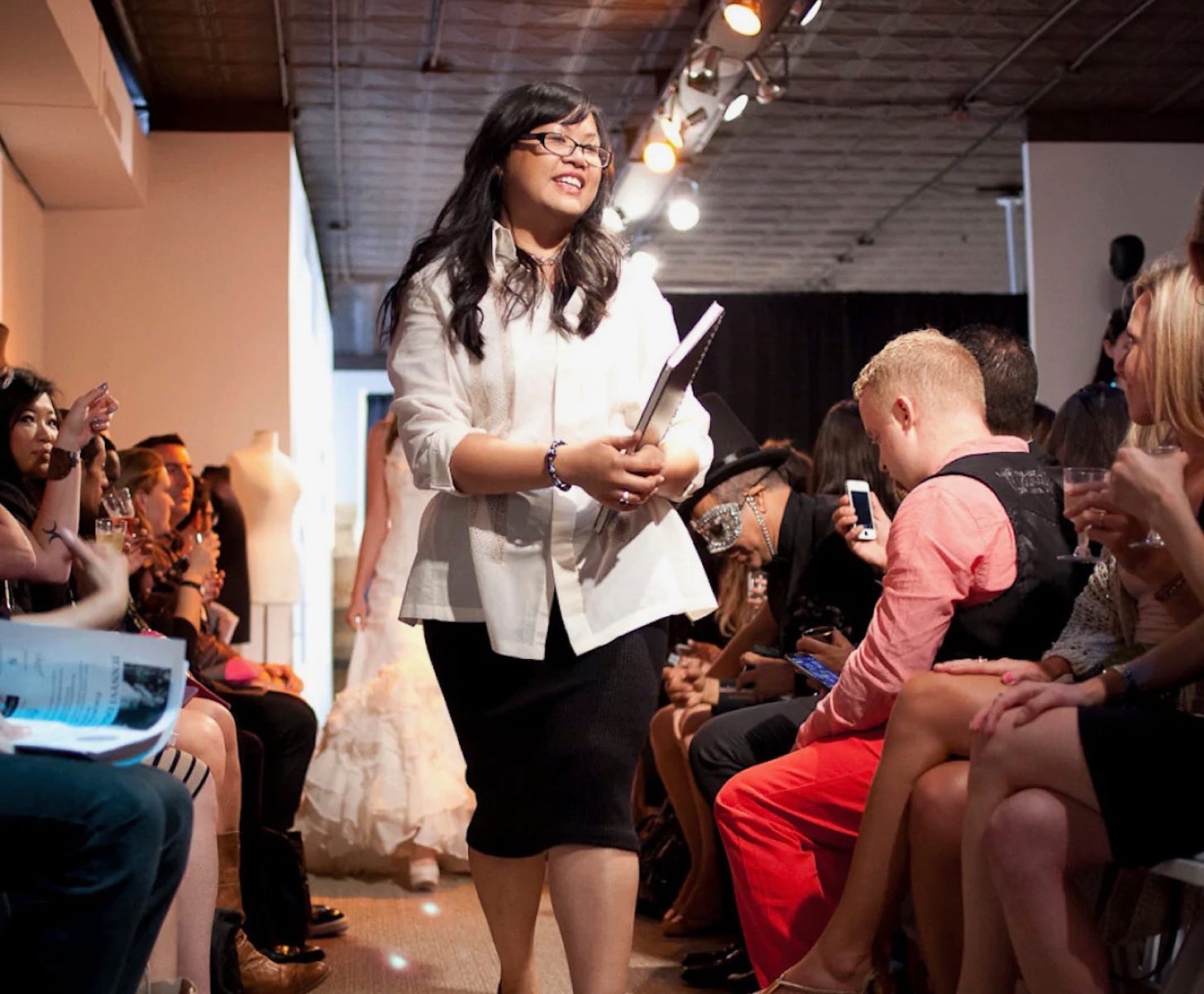 Designer Jennyvi Dizon walks in a very crowded New York Fashion Week show, she's smiling and holding a large book. 