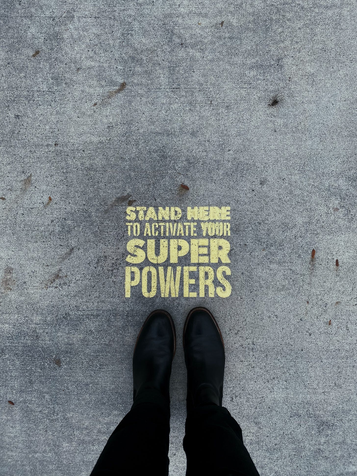 Looking down on feet in black boots on gray pavement with gold letters that say "Stand Here to Activate Your Super Powers!"