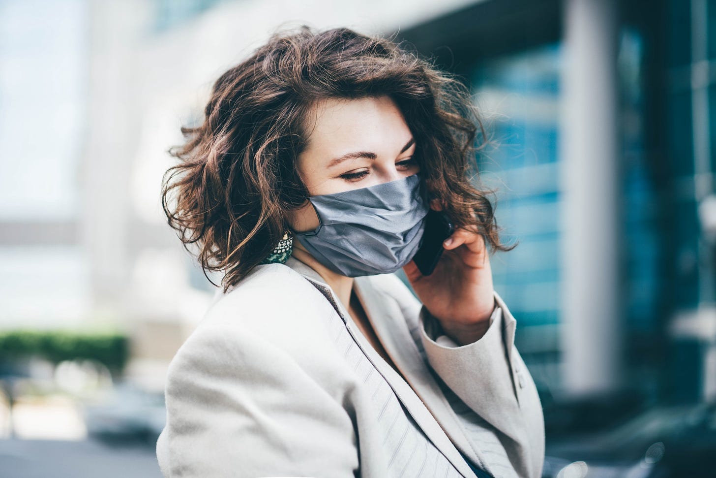 A stylish woman with short brown hair smiles behind a mask in the city.