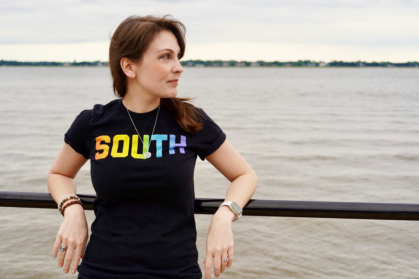 A photo of Kendra, a white woman with brunette hair wearing a black shirt with the word South across the front, standing in front of a vast body of water.
