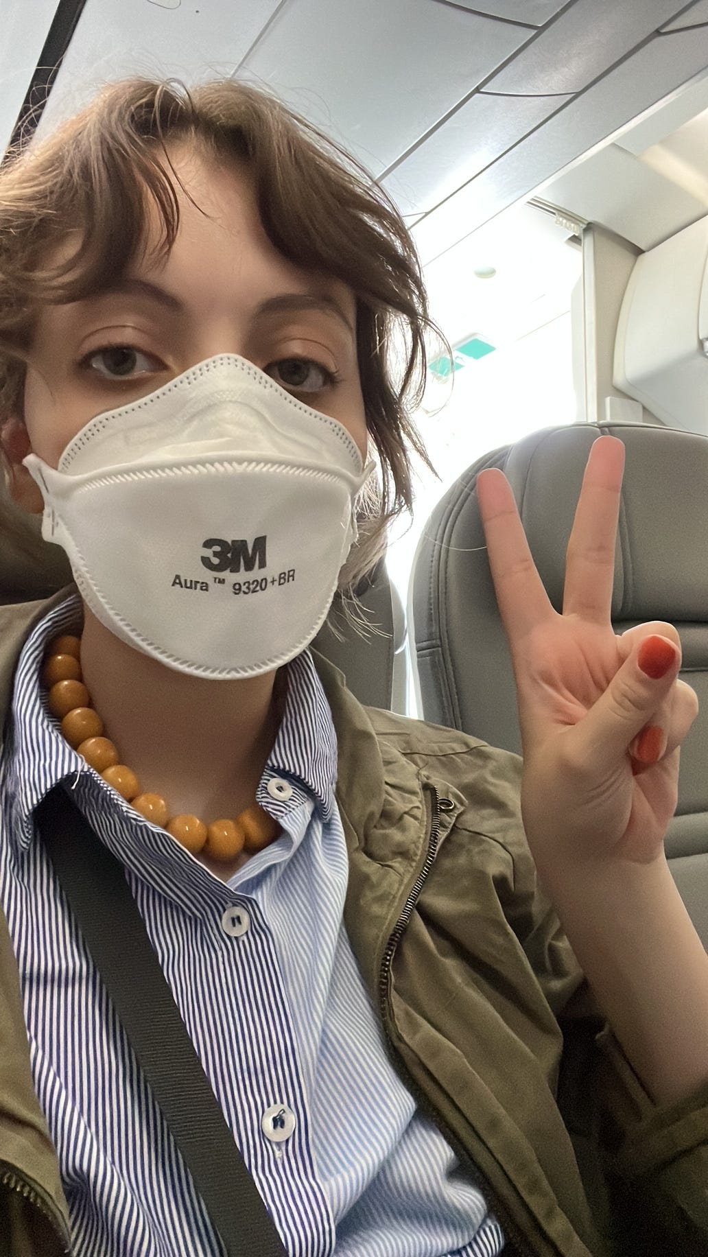 Selfie no avião, fazendo o sinal da paz com os dedos. Estou com os olhos caídos e cara de quem apanhou, cabelo de doida, máscara no rosto, camisa social e casaco verde