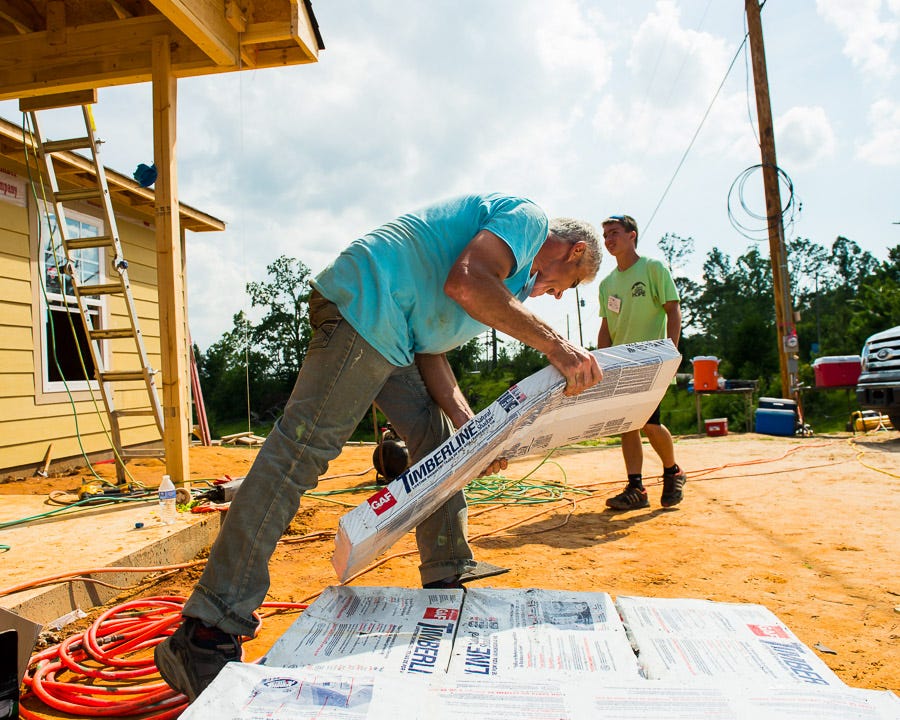 A older man carries shingles as volunteers prepare to replace a roof.