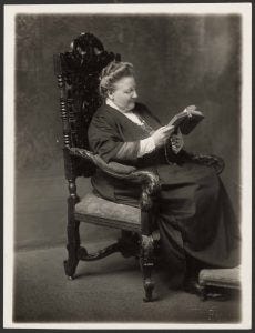 Photo of Amy Lowell seated in a chair reading a book.