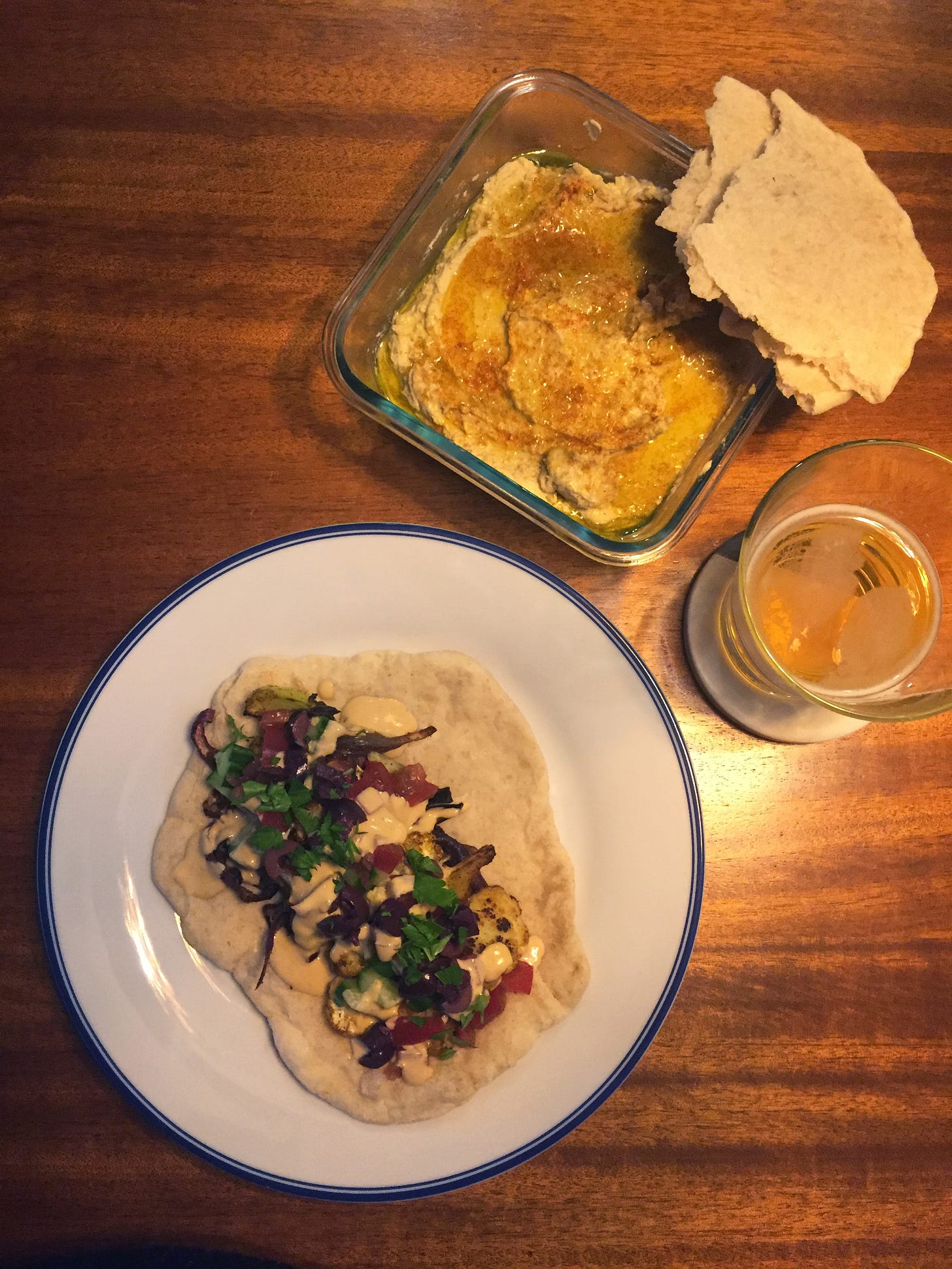 A large plate with a pita covered in roasted cauliflower and chopped vegetables, drizzled with tahini sauce. Above the plate is a square glass container of hummus, coated in olive oil and dusted with paprika and cumin. Torn pieces of pita rest on the side. Next to them on a coaster is a half-full glass of beer.