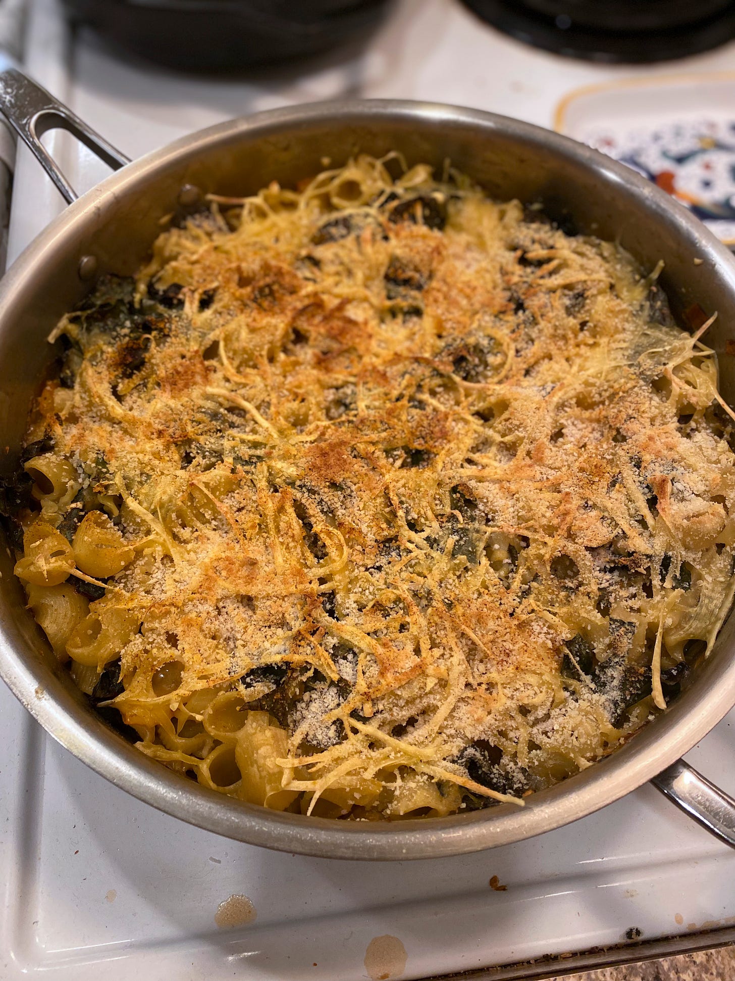 A large stainless steel pan of vegan mac & cheeze with toasty breadcrumbs on top. Pieces of swiss chard are visible throughout.