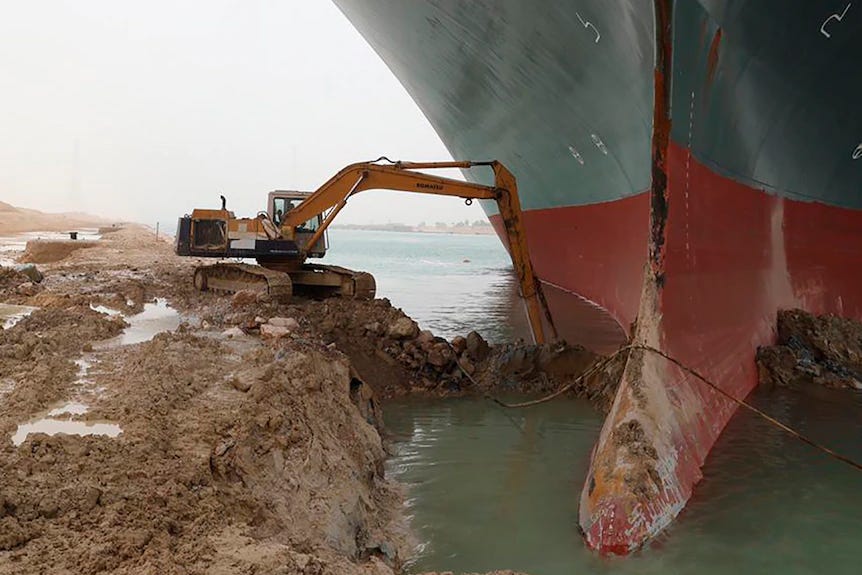An excavator tries to dig out earth from around the bow of a just stupendously large ship lodged in a bank of the Suez Canal.
