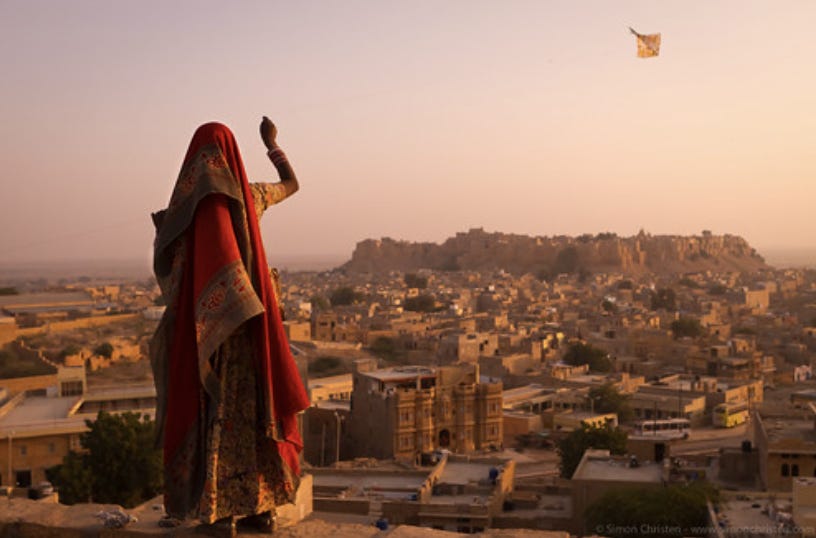 A city filled with golden buildings in the glow of sunset. In the background is a fort on a mountaintop that also looks golden. The sky is golden pink. In the foreground of the picture stands a woman wearing traditional red sari with her back to the camera flying an orange kite. The kite is in the sky to the top right of the sky.