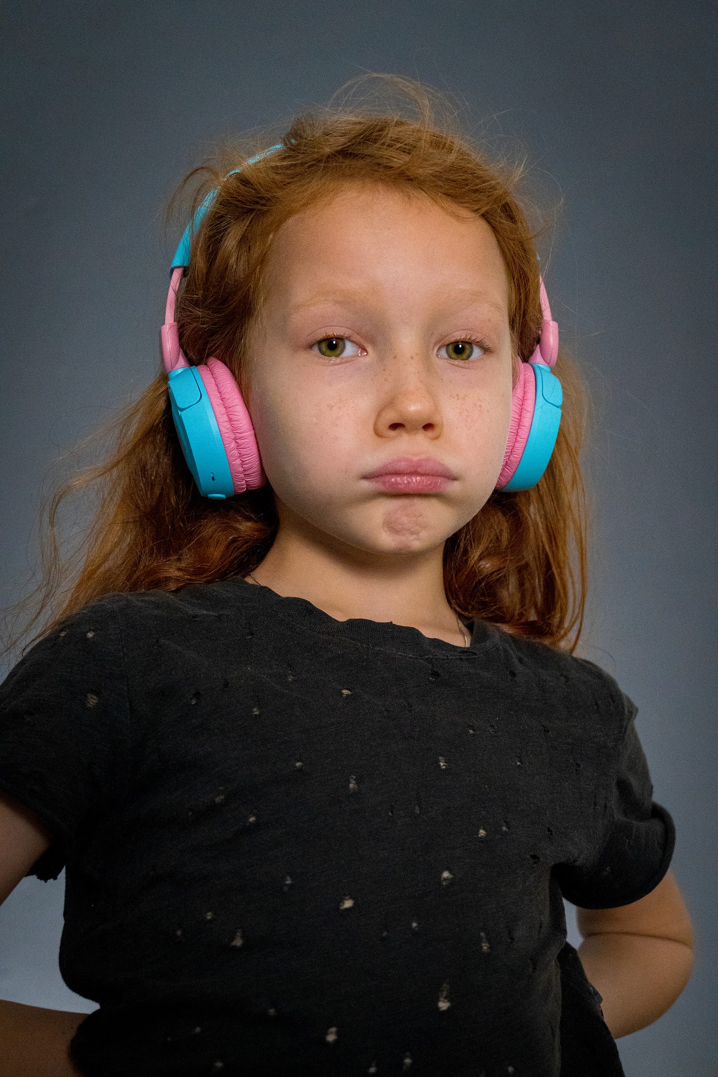  a red haired girl is reading a black shirt and bright pink and blue headphones. She is blowing her cheeks out in disapproval. Her hands are on her hips.
