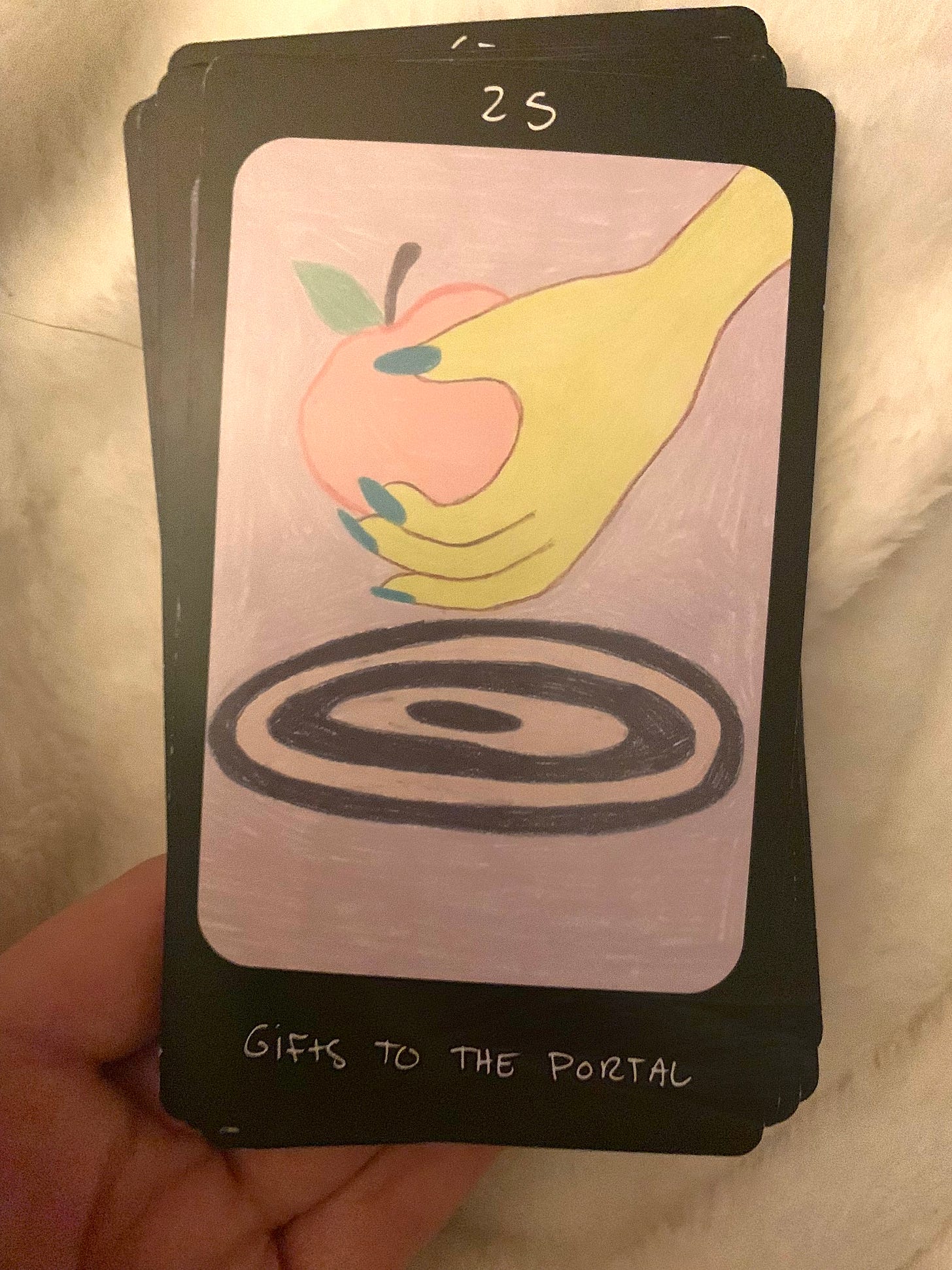 mira's left hand holds a stack of oracle cards over a fuzzy white blanket. the top card reads, "25" on the top and "GIFTS TO THE PORTAL" on the bottom. the card has a black border, a lavender background, and depicts an image of a hand offering an apple to a portal.
