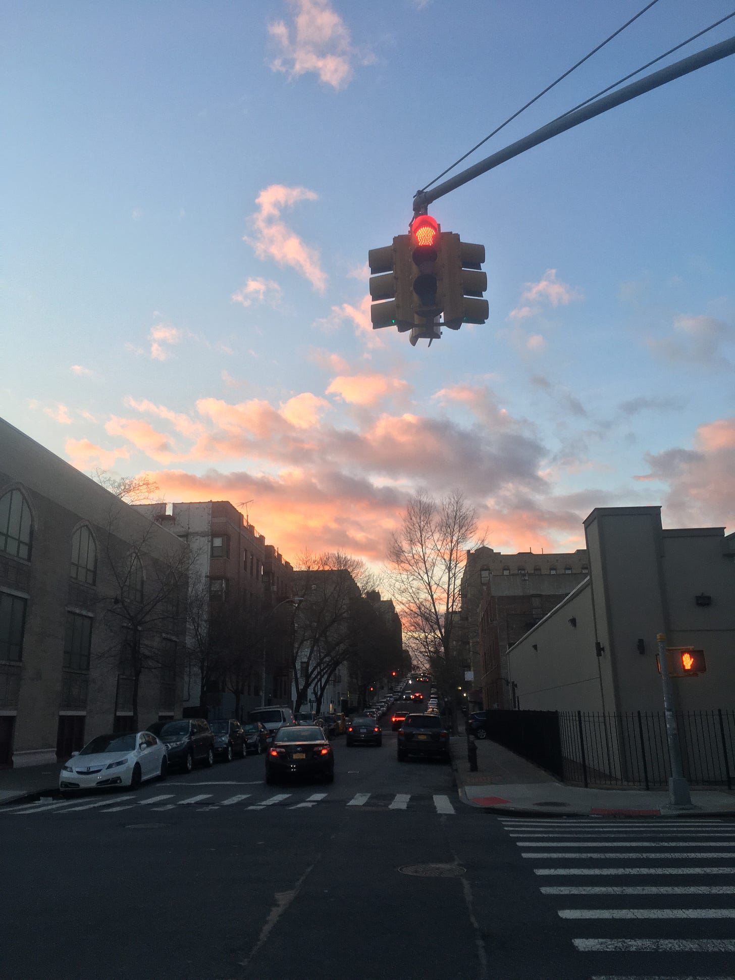 Photo by me: pink clouds at the bottom of a mostly clear sky. Below, cars in the street and a stop light at red
