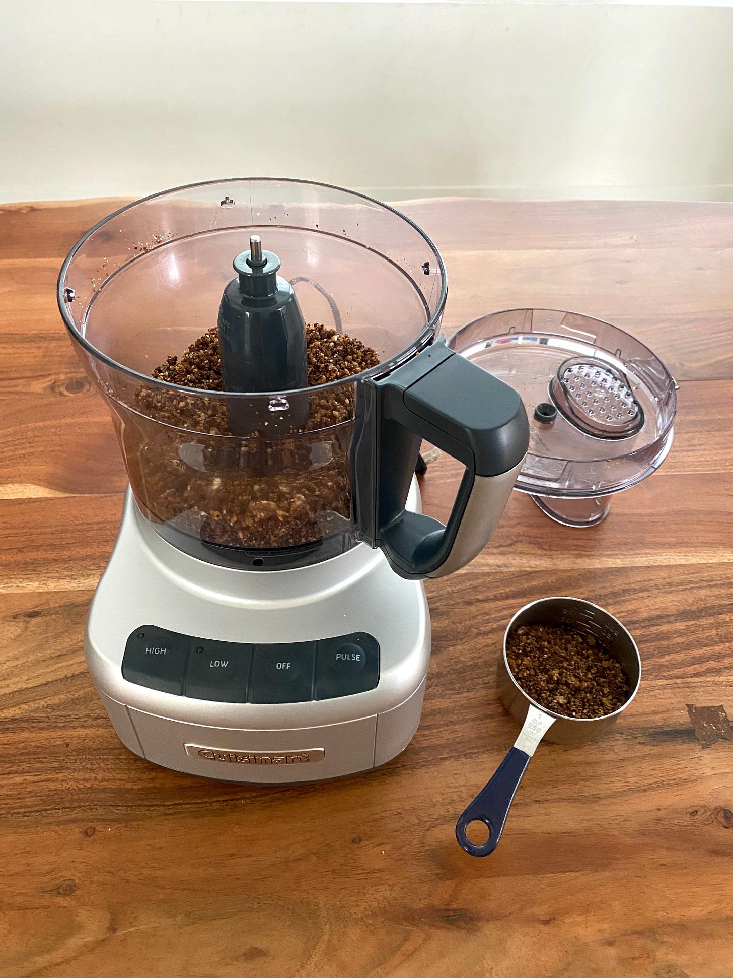 An open food processor filled with chocolate cookie crumbs rests on a wooden table. The lid and a measuring cup half-filled with cookie crumbs rests next to it.