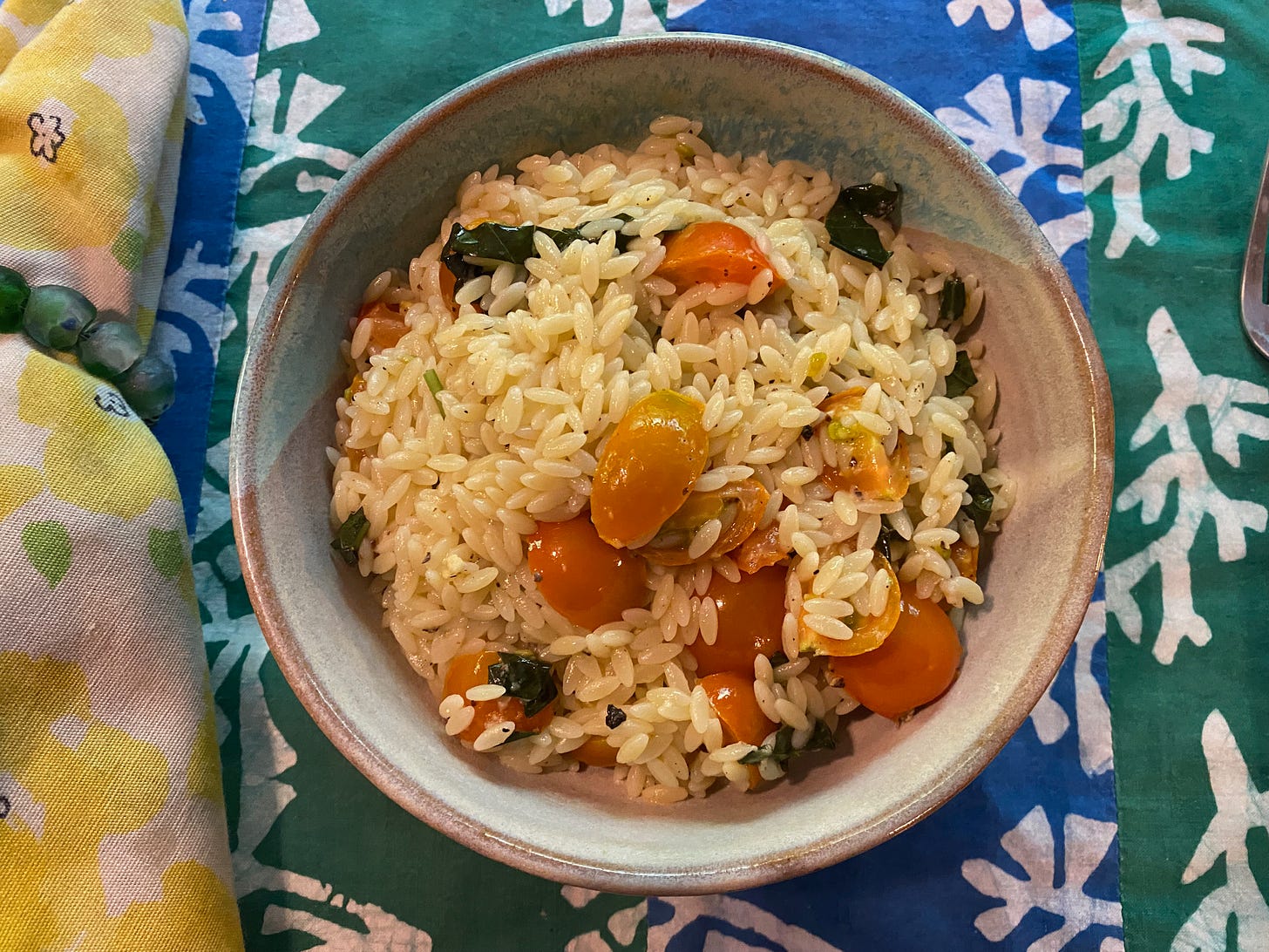 A ceramic bowl of orzo, halved sungold cherry tomatoes, and chopped basil sits on a blue and green placemat next to a yellow cloth napkin.