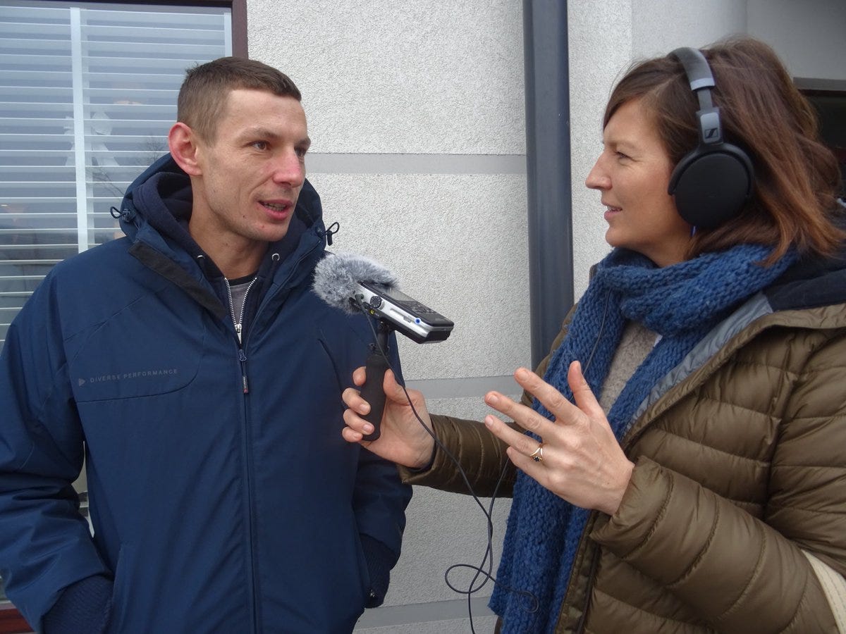 foto van Maartje die met haar recorder in hand en koptelefoon op Pawel, een witte man met kort haar en een blauwe jas, op straat interviewt.