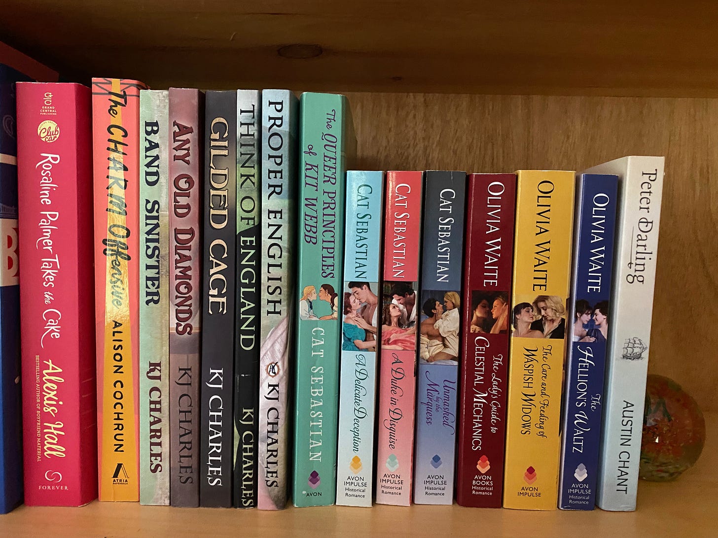 A close-up of a shelf of queer romance novels, trade paperbacks and mass markets. The books are: Rosaline Palmer Takes the Cake by Alexis Hall, The Charm Offensive by Allison Cochrun, Band Sinister, Any Old Diamonds, Gilded Cage, Think of England, and Proper English by KJ Charles, The Queer Principles of Kit Webb, A Deliciate Deception, A Duke in Disguise, and Unmasked by the Marquess by Cat Sebastian, the Feminine Pursuits trilogy by Olivia Waite, and Peter Darling by Austin Chant.