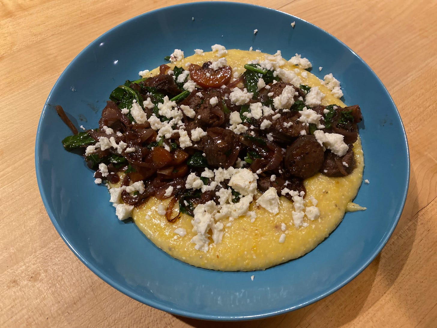 A wide blue bowl full of creamy polenta topped with kale, pieces of sausage, carrots, and crumbled feta cheese.