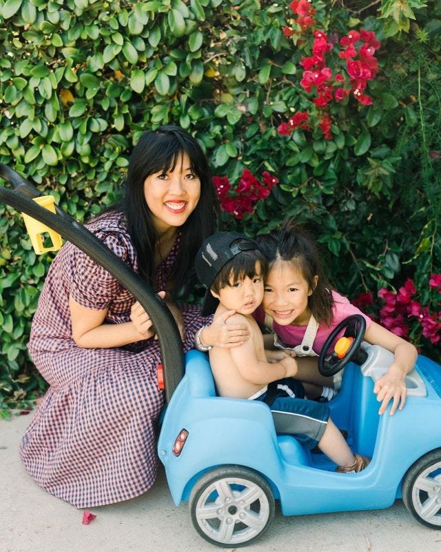 Michelle, an Asian American woman, crouches next to her two children, who are playing on a drivable toy car, against a background of green leaves and pink flowers. 