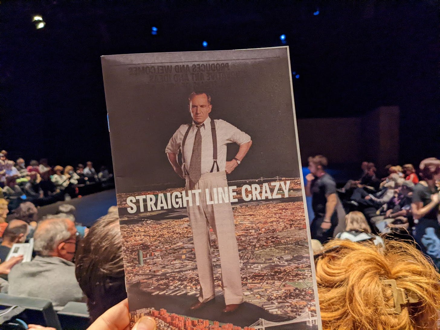 A hand holds a playbill that reads "Straight Line Crazy." Behind the playbill is an audience seated around a stage.
