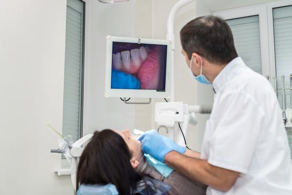 A imagem contém um dentista fazendo um exame intraoral, tecnologia da Odontologia Digital, na boca da paciente. Na tela aparecem os dentes da paciente.