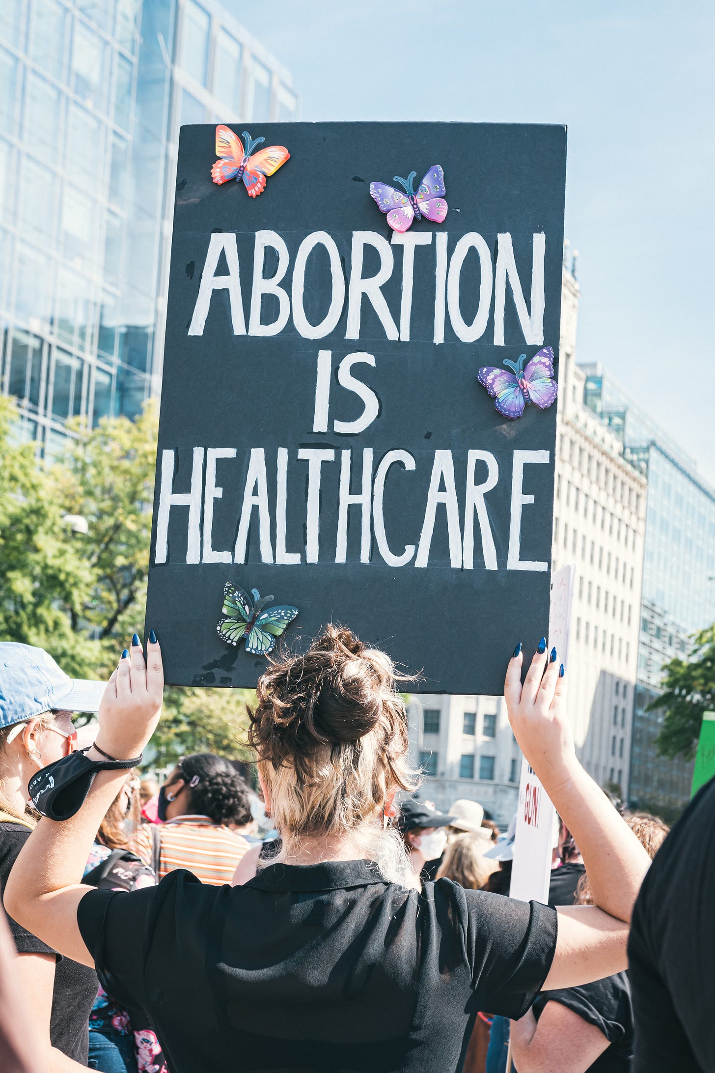 A close-up of a homemade sign at an abortion rights protest. On a black background, text in white lettering says ‘Abortion Is Healthcare’. The slogan is surrounded by butterflies.