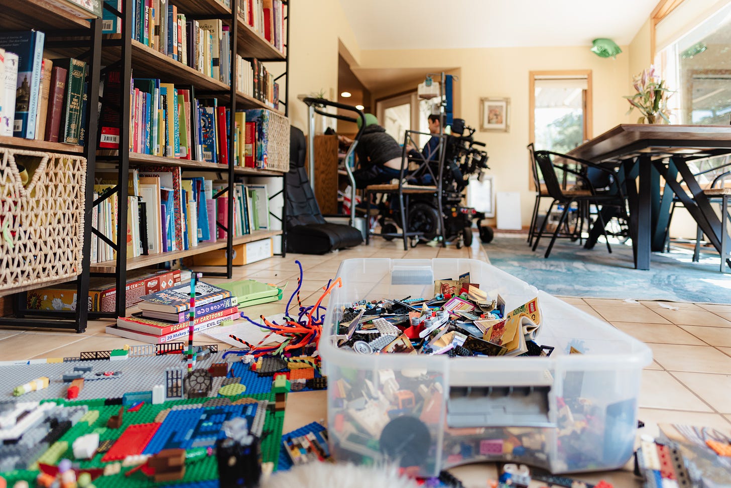 In the foreground, a pile of LEGO and other toys. Along one wall are full bookshelves. In the background, a boy in a wheelchair and another on a dining chair crowd next to a smart speaker. 