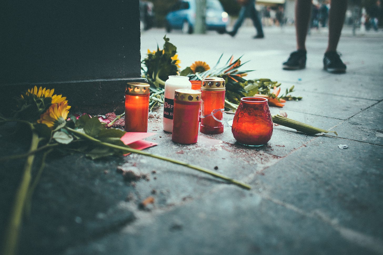 Candle and flowers on the ground in a public street as people walk by and one person stands by them Candle and flowers on the ground in a public street as people walk by and one person stands by them